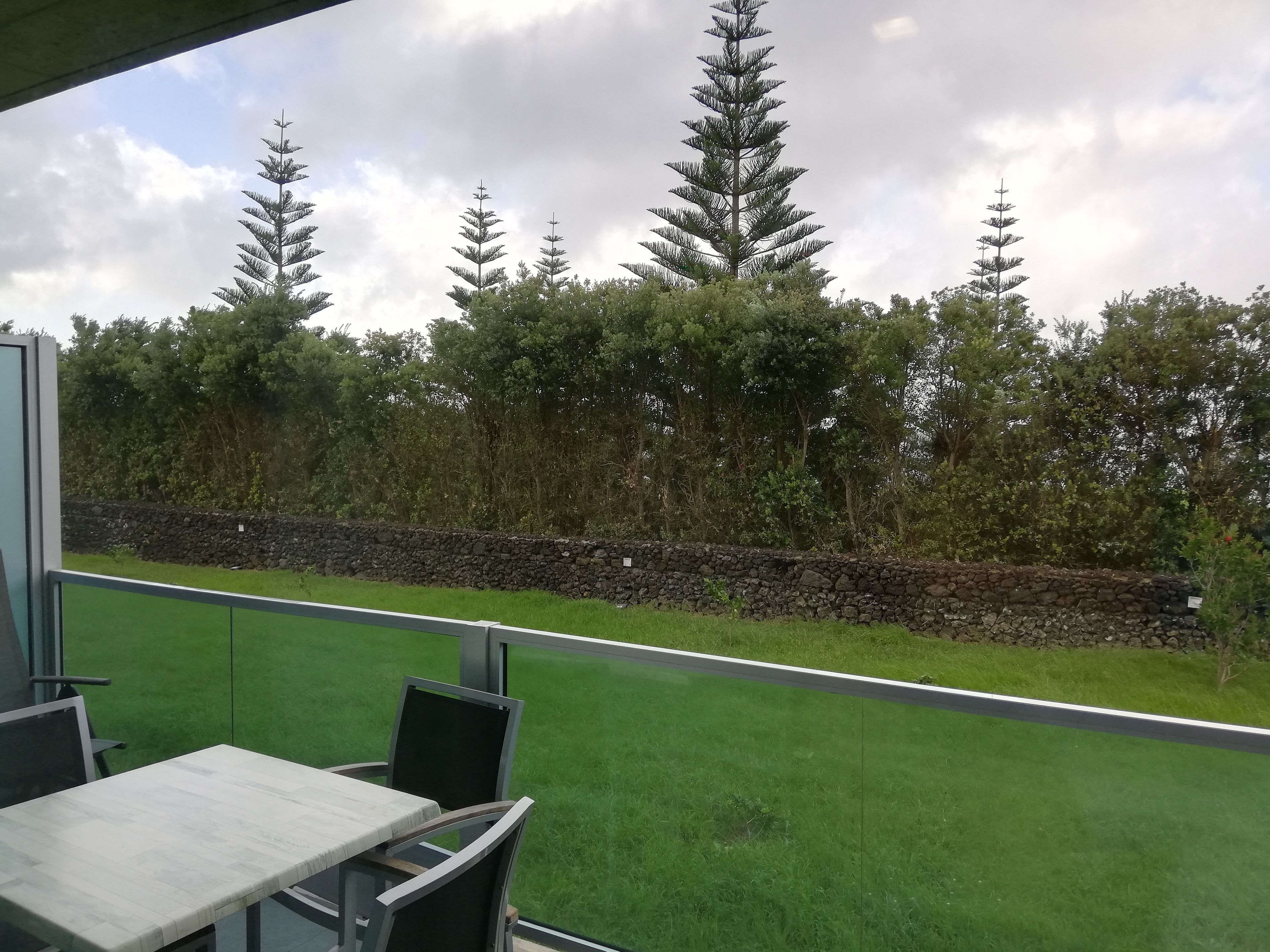 Ein Balkon mit Tisch und Stühlen, der auf eine Rasenfläche mit einer Steinmauer und Bäumen blickt.