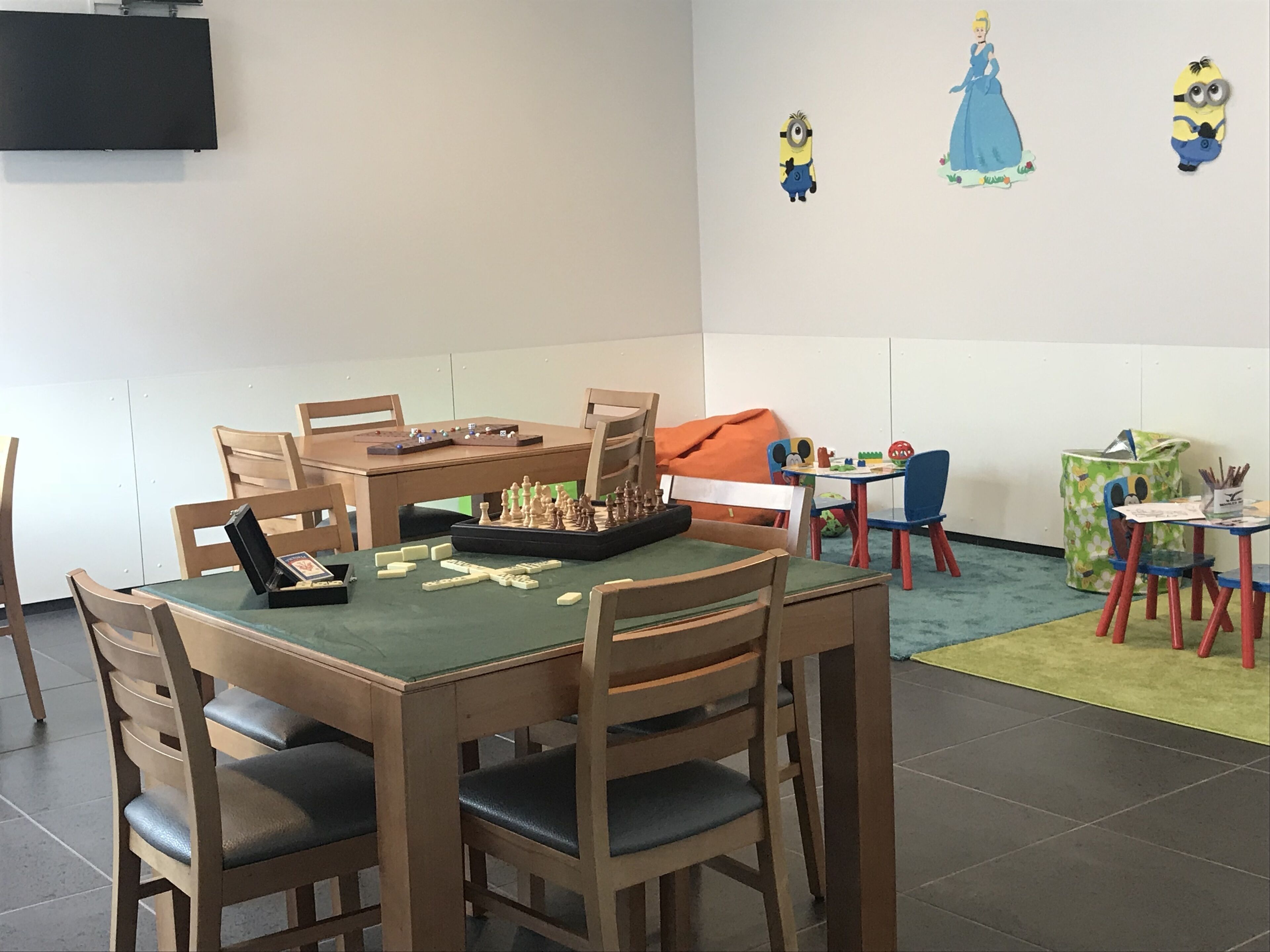 Una habitación con un tablero de ajedrez sobre una mesa, sillas de madera, un televisor en la pared y una decoración de pared con temática de los Minions.
