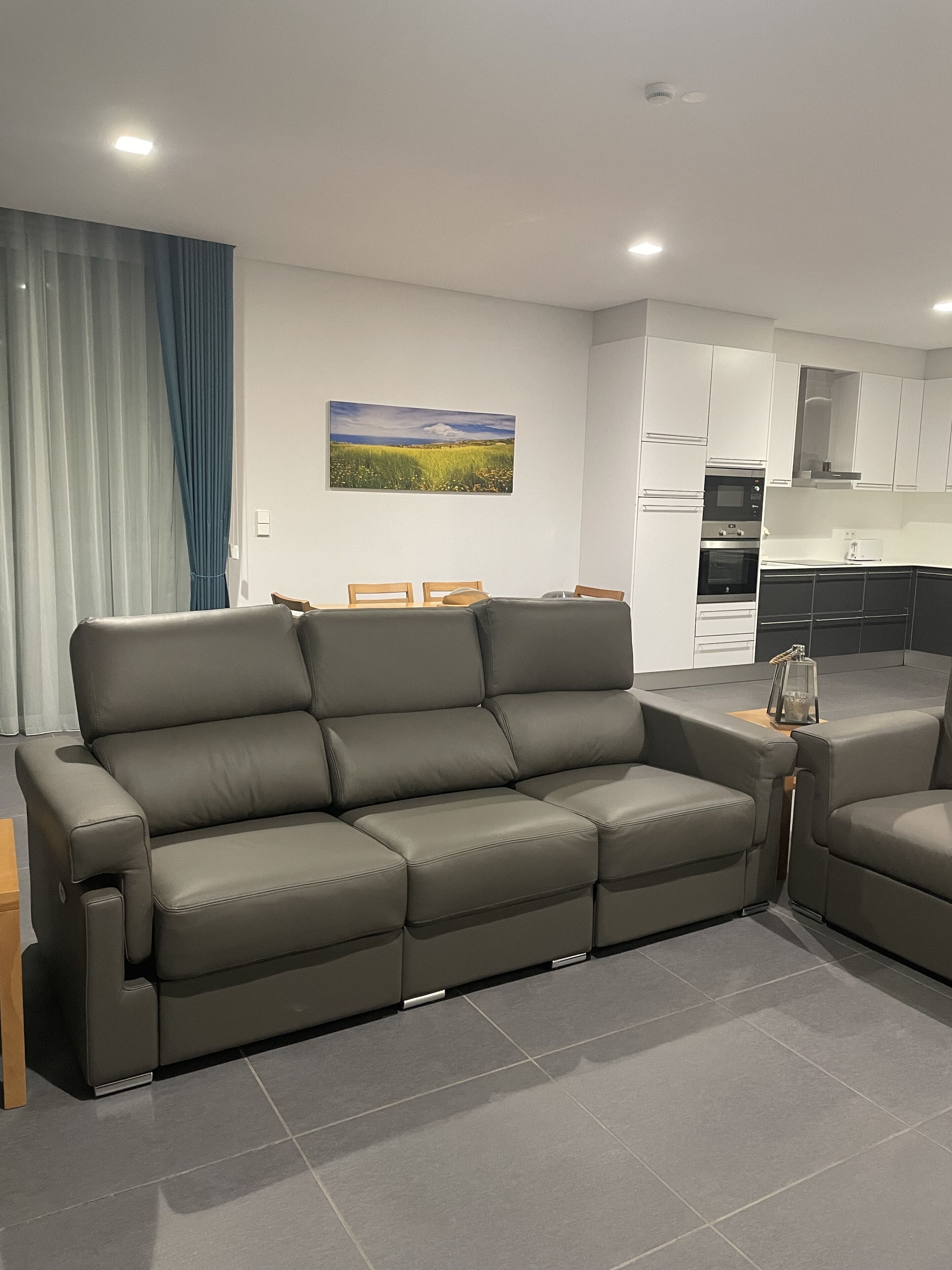Una sala moderna con un sofá seccional gris, un comedor con mesa y sillas de madera, y una cocina con gabinetes y electrodomésticos blancos.