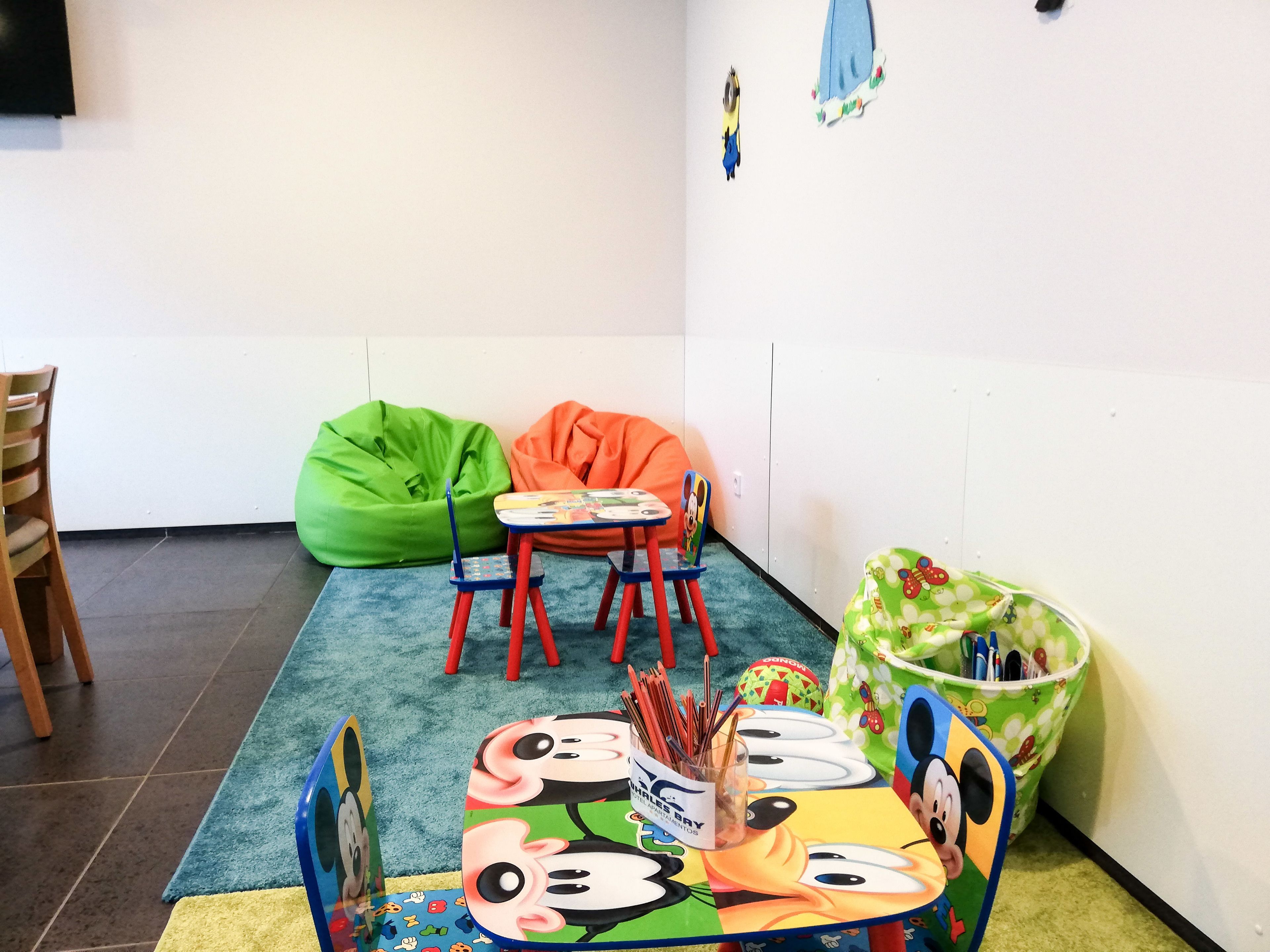 Un espace de jeux pour enfants avec des poufs colorés, une petite table recouverte d’une nappe à l’effigie de Mickey Mouse et un sac vert orné d’un motif de papillon.