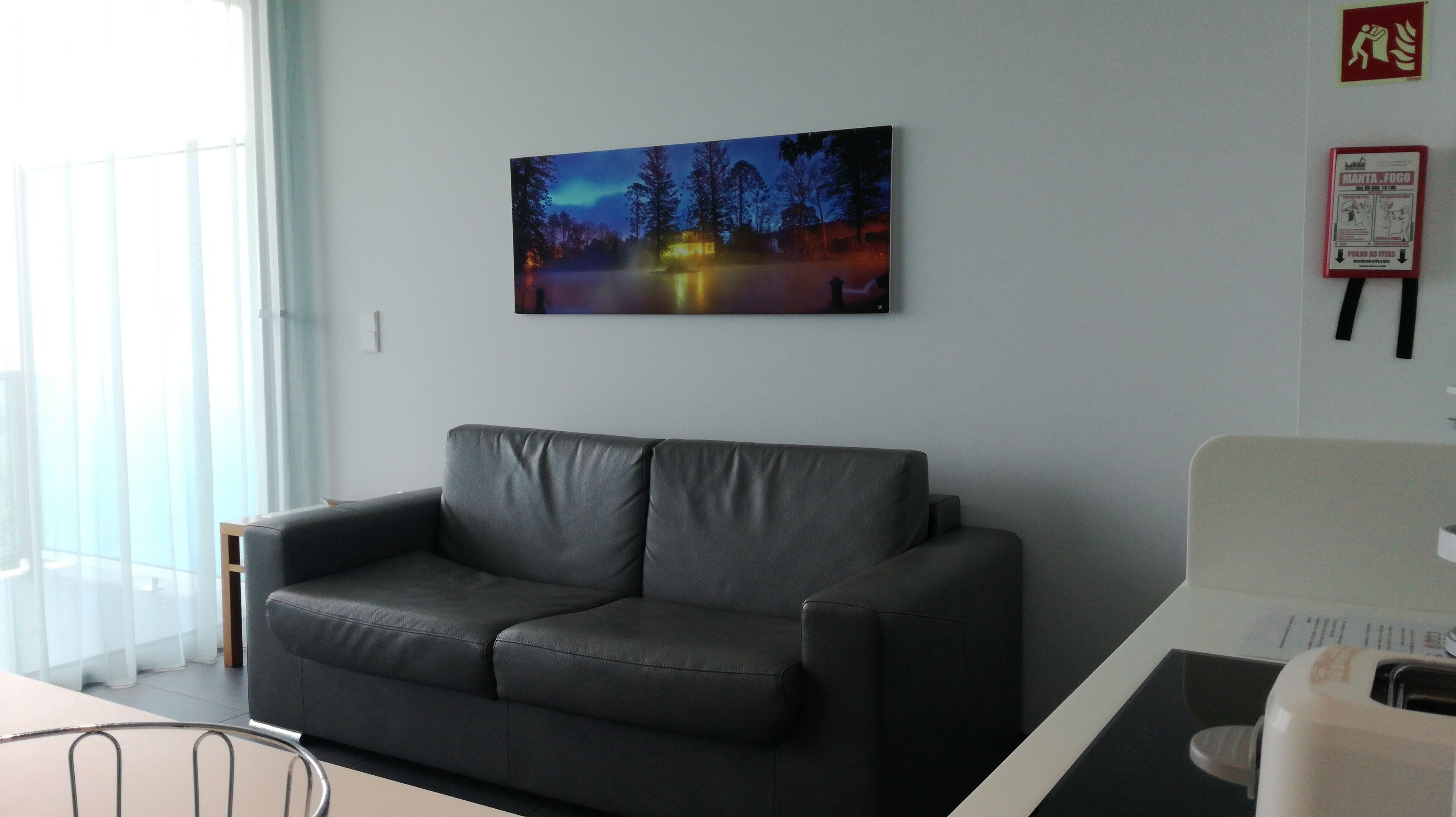 A living room with a dark leather sofa, a flat-screen TV mounted on the wall, and a window with sheer curtains.
