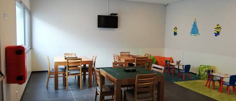 A dining area with wooden tables and chairs, a television mounted on the wall, and a children's play area with colorful decorations.