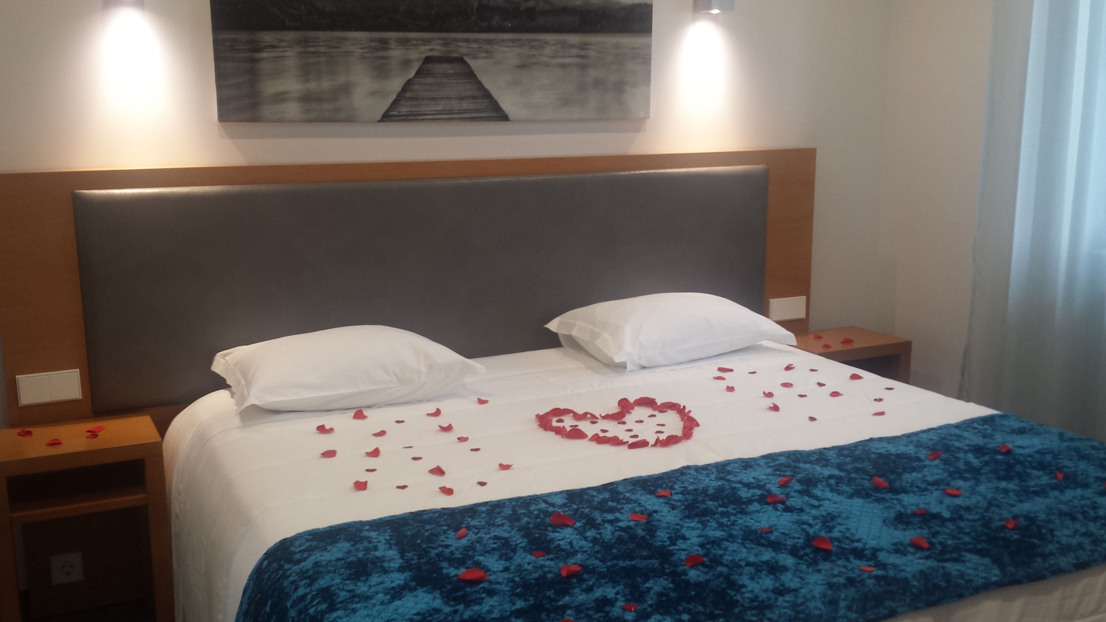 Ett hotellrum med en stor säng, två vita kuddar, ett blått täcke och röda rosenblad som är utspridda på sängen.
