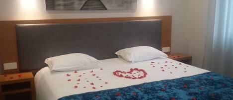 Ett hotellrum med en stor säng, två vita kuddar, ett blått täcke och röda rosenblad som är utspridda på sängen.