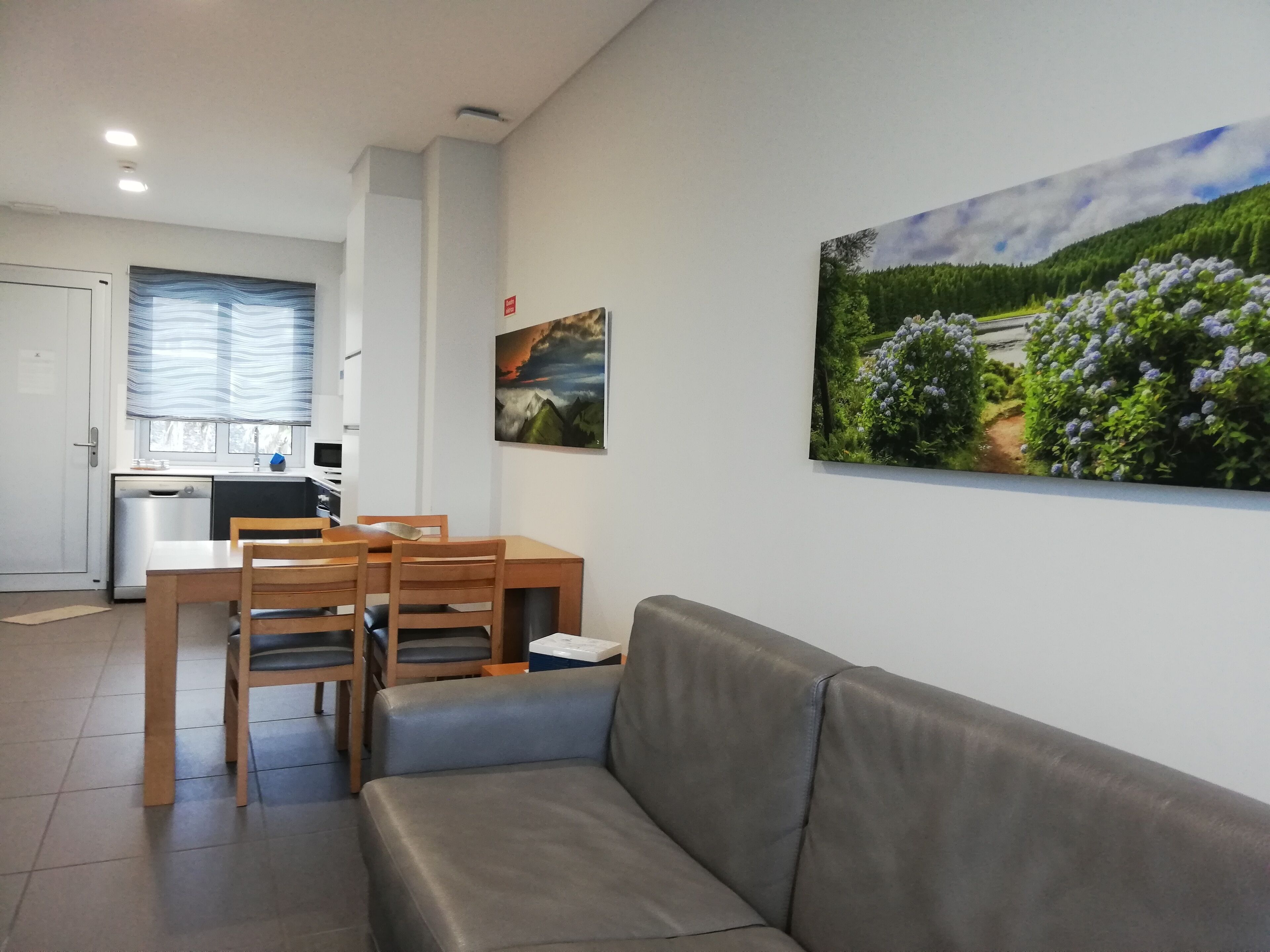 Una sala de estar moderna con un sofá gris, una mesa de comedor de madera y sillas. Se ve una cocina con lavavajillas y una ventana con persianas en el fondo.