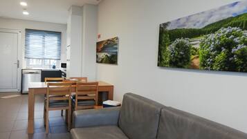 Una sala de estar moderna con un sofá gris, una mesa de comedor de madera y sillas. Se ve una cocina con lavavajillas y una ventana con persianas en el fondo.