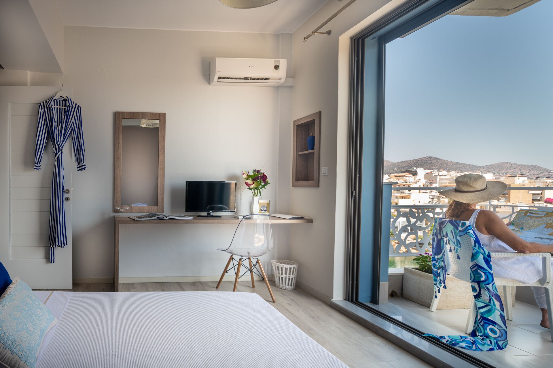 Une chambre d’hôtel moderne dotée d’un grand balcon, d’un lit, d’un bureau avec un ordinateur et offrant une vue sur le paysage urbain.