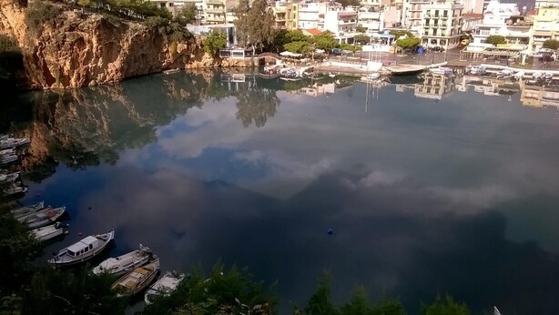 A calm lake with boats docked on the left, surrounded by buildings and greenery. - Emerald Lake Studios (Agios Nikolaos)