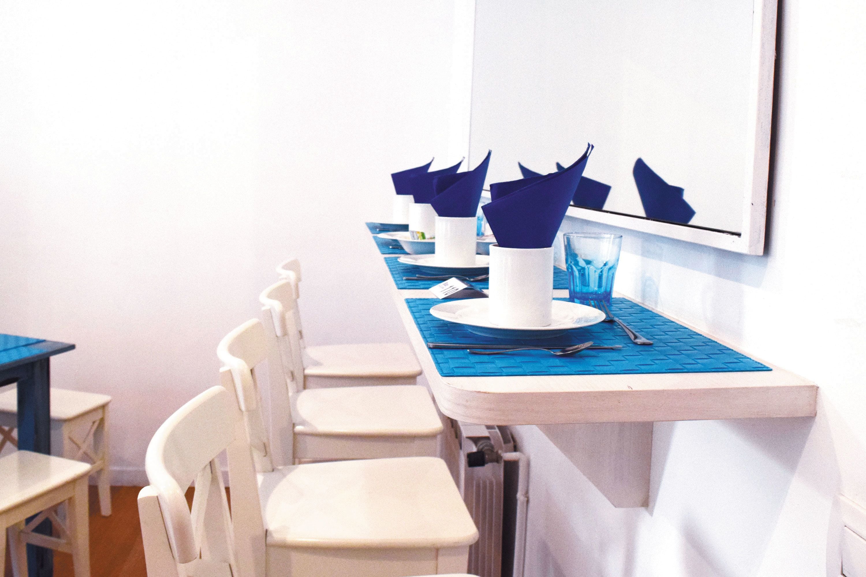 Un espace repas avec des chaises et des tables blanches, des sets de table bleus et des assiettes blanches accompagnées de serviettes bleues.