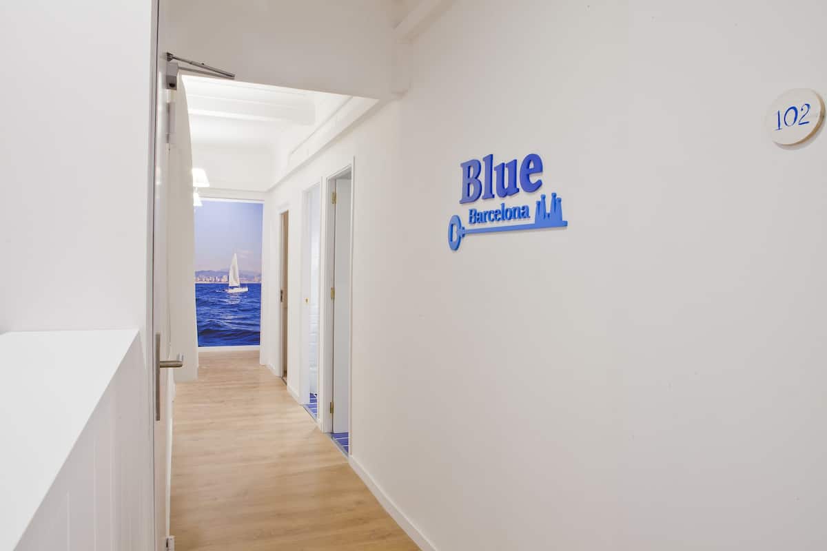 A hallway with a wooden floor leading to a room with a view of the sea. A sign on the wall reads "Blue Barcelona."