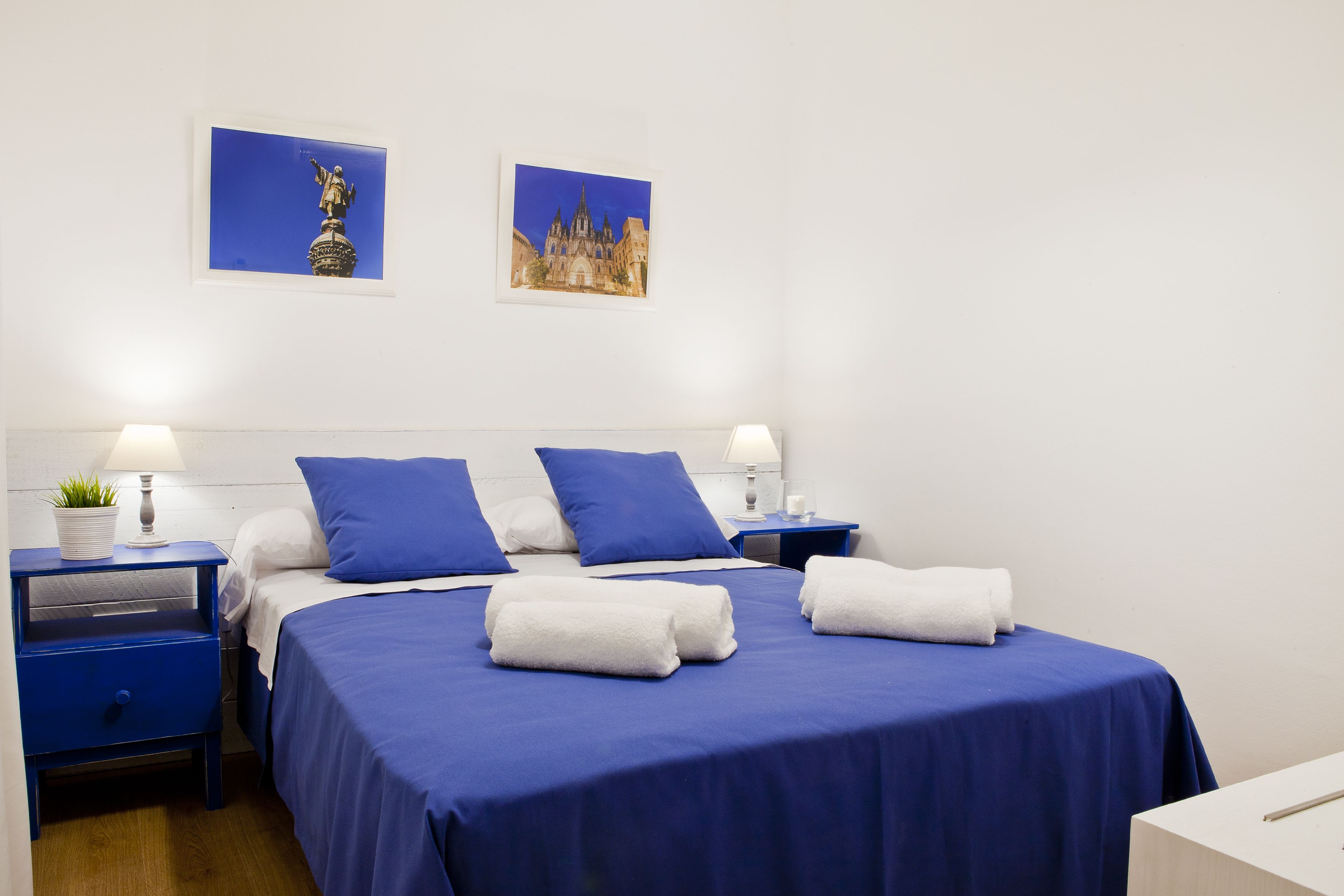 Habitación de hotel con una cama grande, dos mesitas de noche, una lámpara, una planta en maceta y dos cuadros en la pared.