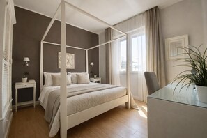 A four-poster bed with a white frame, two bedside tables with lamps, a glass desk, and a potted plant.