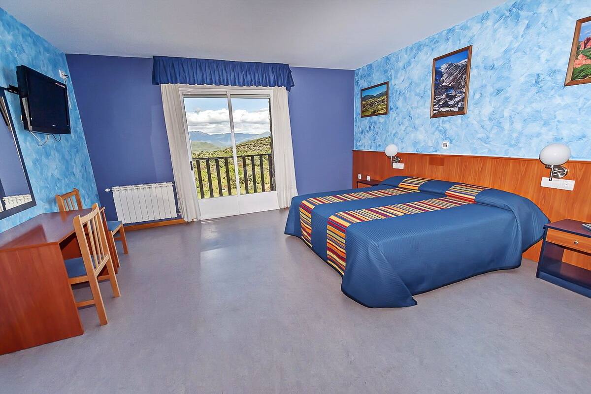 A bedroom with two beds, a desk, and a chair. There is a window with a view of a balcony.