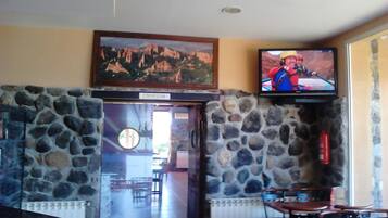Un restaurante con paredes de piedra, un cuadro colgado y una pantalla de televisión que muestra a una persona con chaqueta roja.