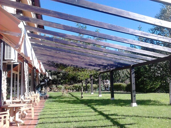 A covered outdoor seating area with tables and chairs, a grassy lawn, and a building with a pergola in the background. - Hotel Medulio (Carucedo)