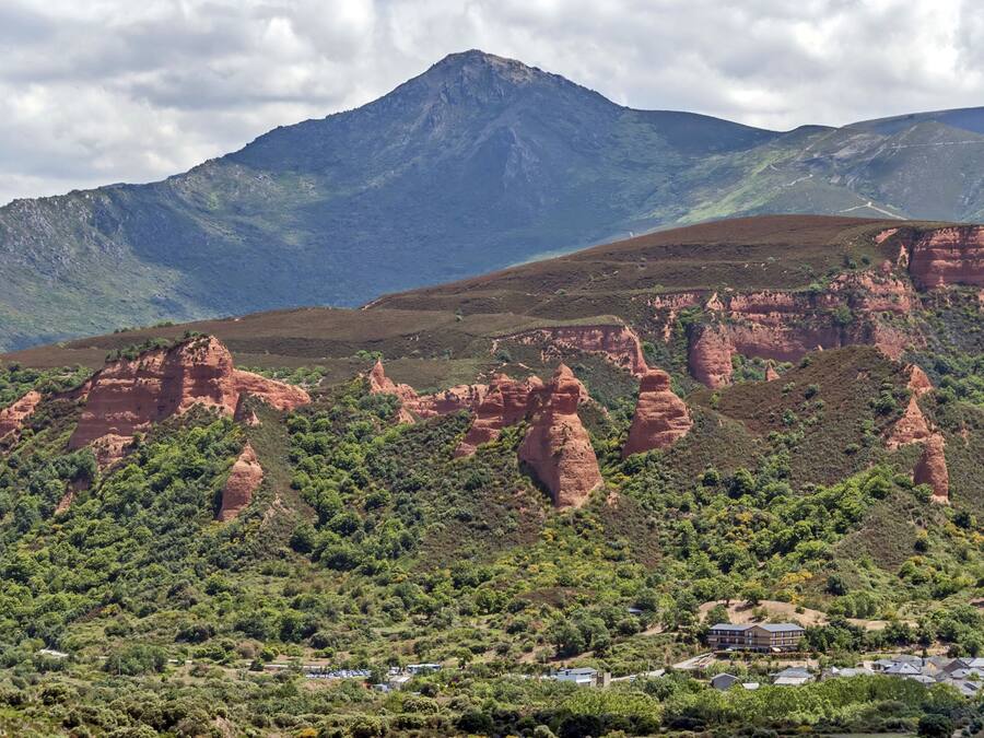 A mountainous landscape with a prominent peak, red rock formations, and a small cluster of buildings at the base.