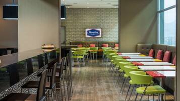 A hotel dining area with a long counter, green and red chairs, and a stone wall with a 'Hampton by Hilton' sign.