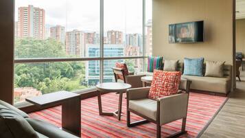 A modern hotel room with a large window offering a city view, a sofa, a chair, a small table, and a TV mounted on the wall.