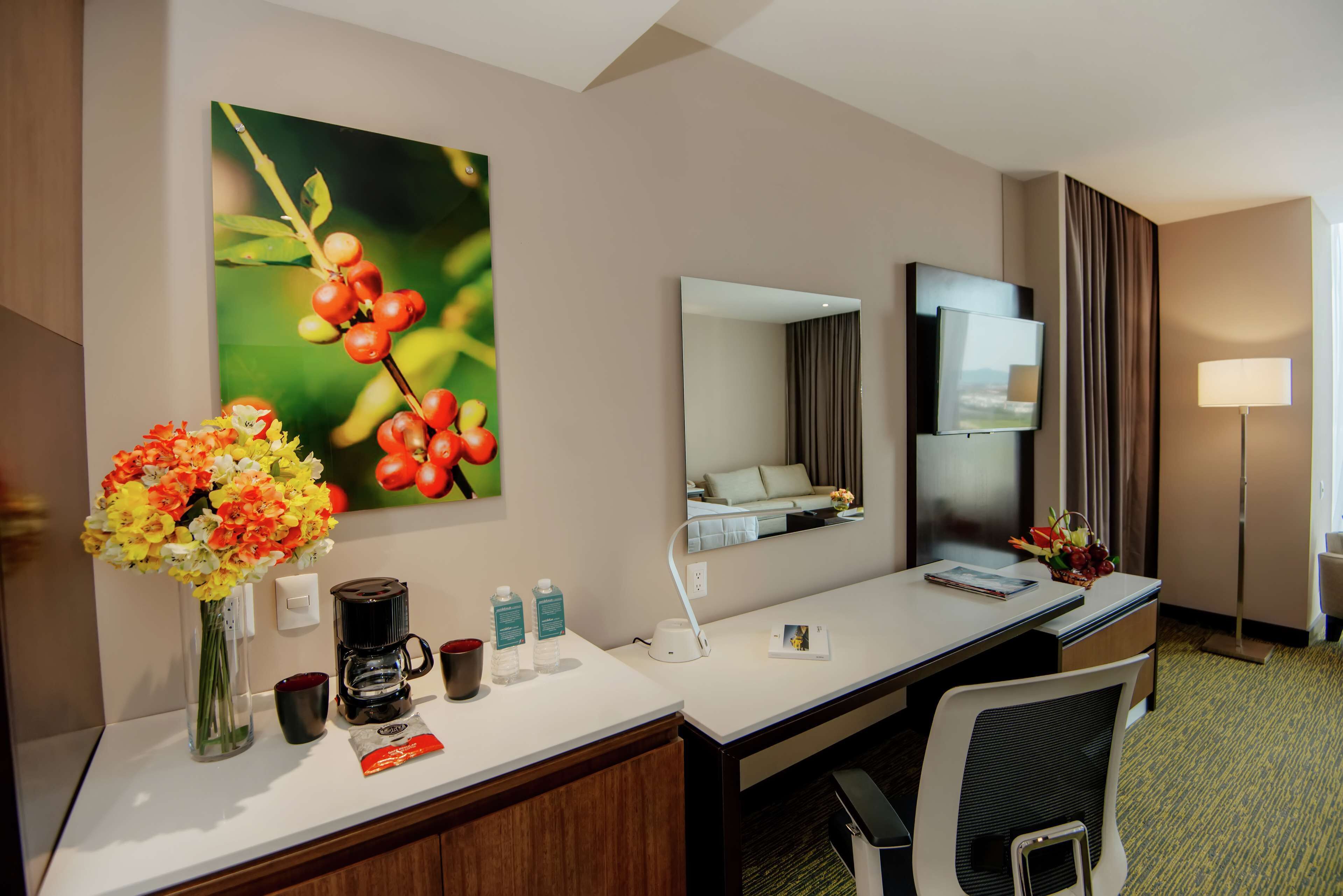 A hotel room with a desk, a large mirror, a coffee maker, a vase with flowers, and a painting of berries.