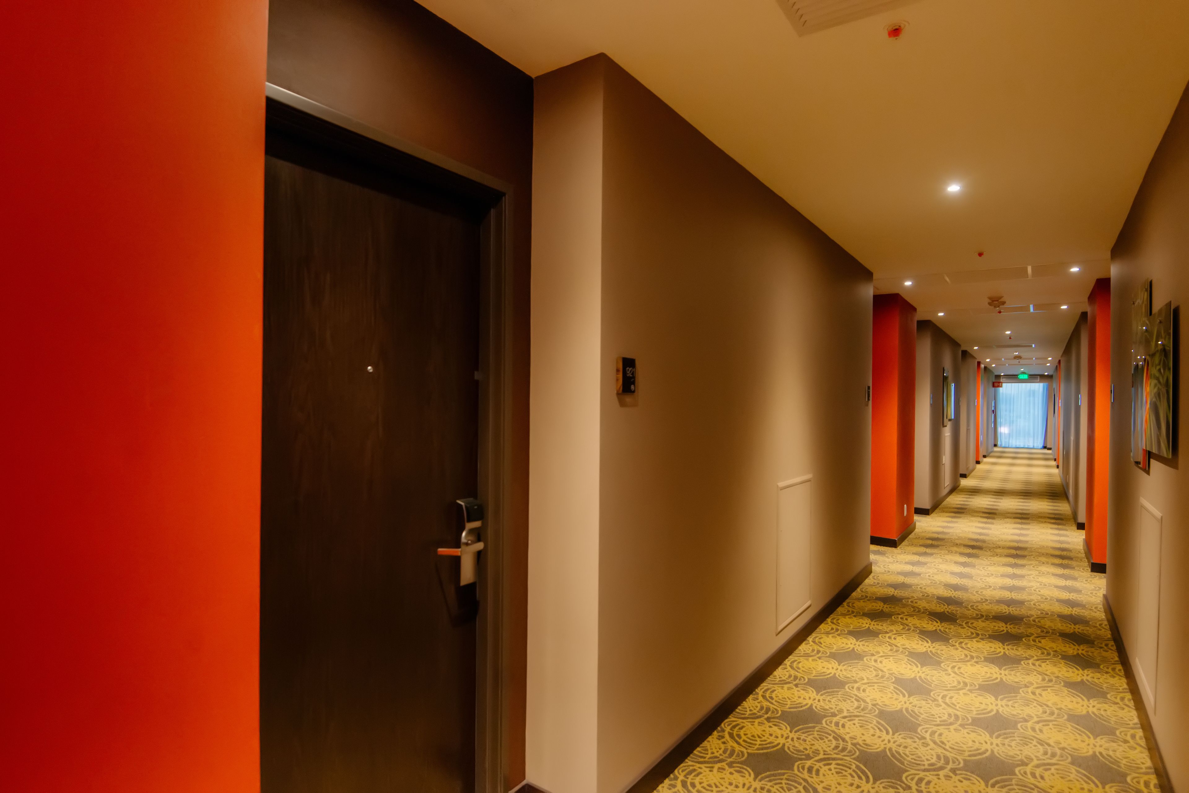 Ein Hotel-Flur mit einem gemusterten Teppich, einer Holztür und einem Gang, der in einen hellen Raum führt.