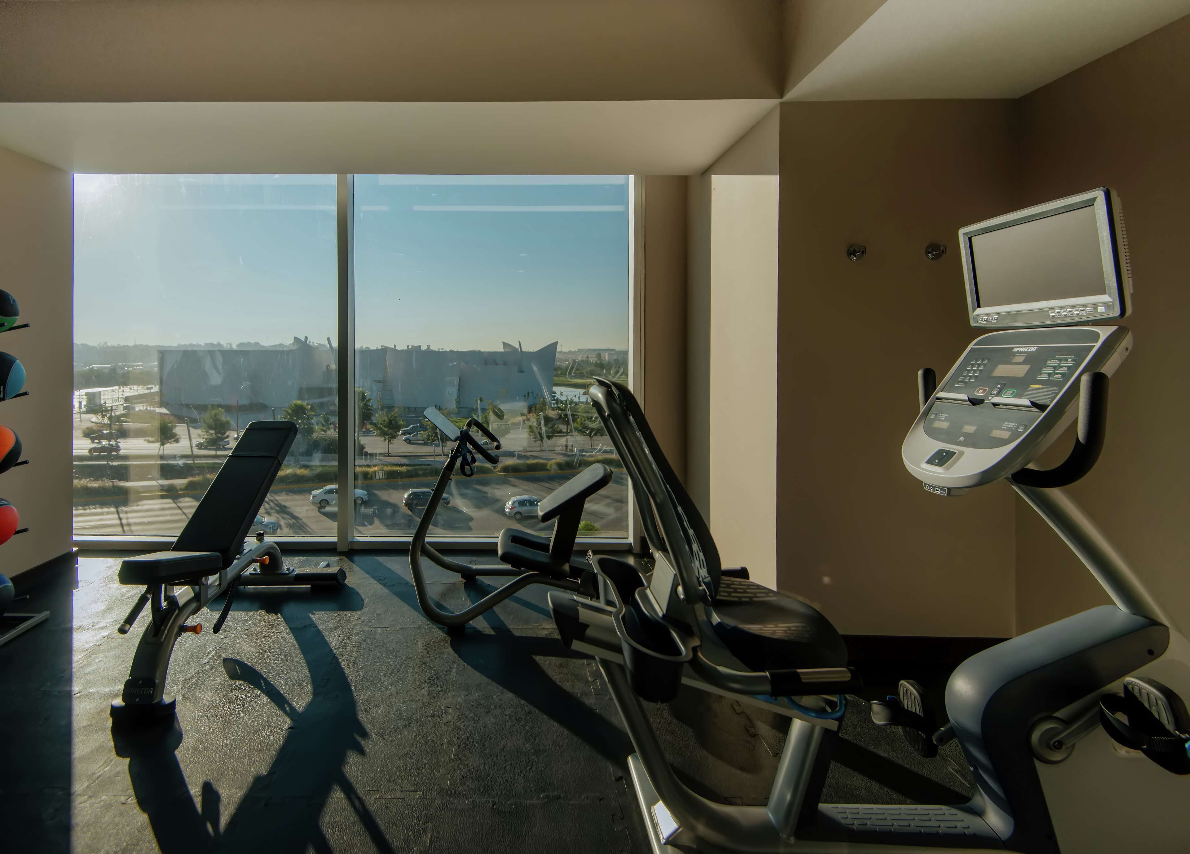 A gym with exercise equipment, large windows, and a view of a parking lot and buildings.