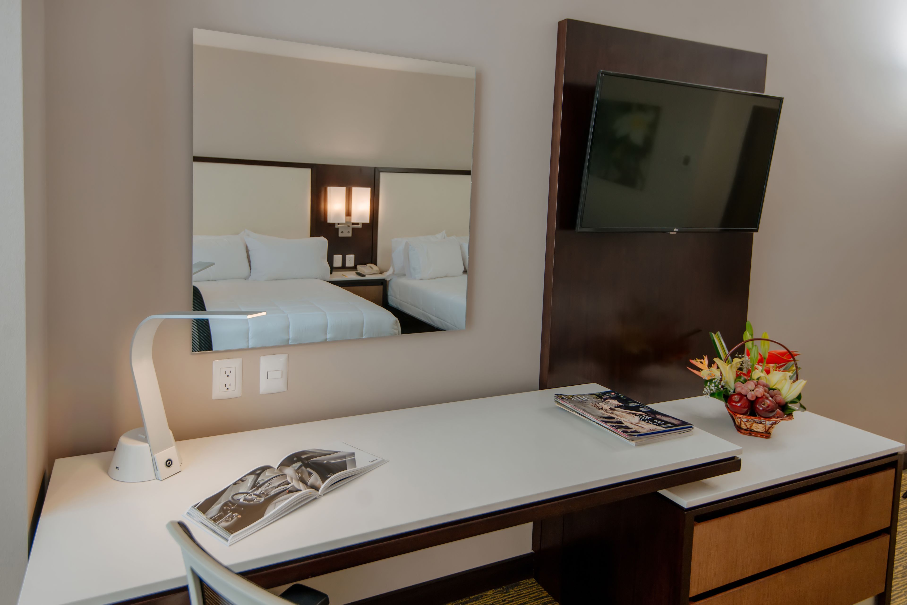 A hotel room with a bed, bedside table with a lamp, a TV mounted on the wall, and a small table with a magazine and a fruit basket.