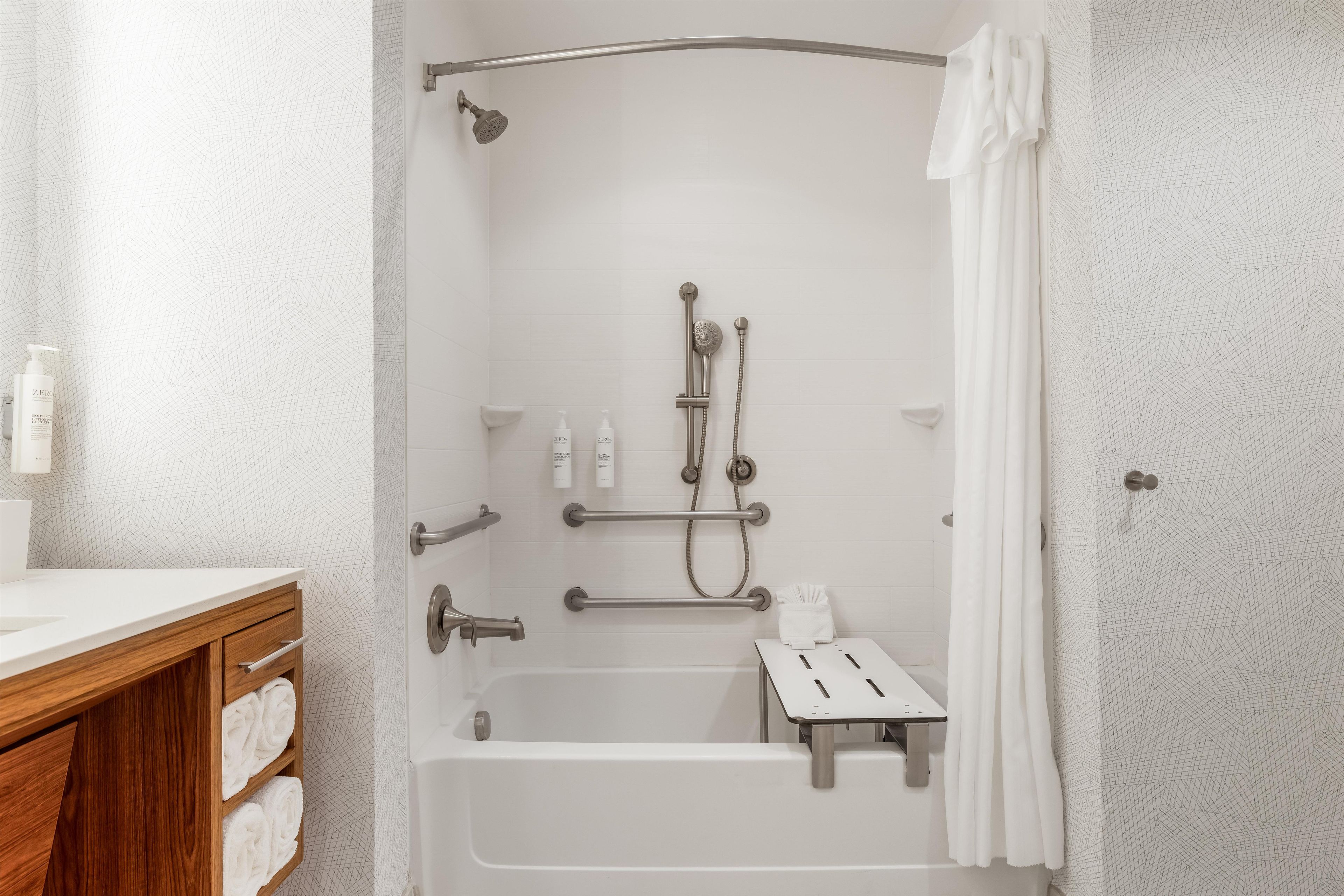 Un baño moderno con ducha a nivel, barras de apoyo, una bañera blanca y un mueble de tocador de madera con toallas.