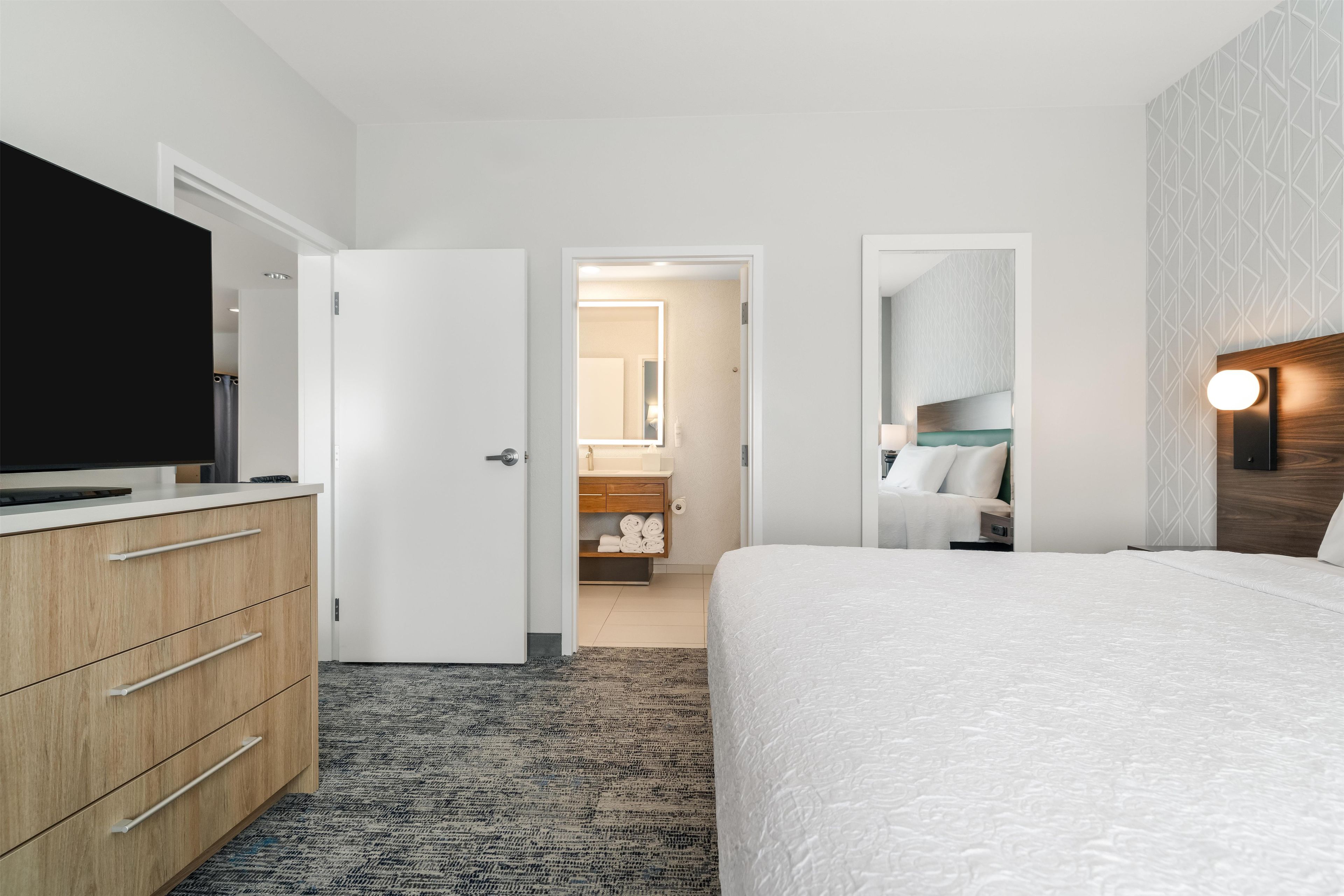 A hotel room with a bed, a TV, a dresser, a bathroom visible through an open door, and a patterned wall.