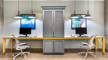 A modern office space with two desks, each with a computer monitor, keyboard, and phone. There are two office chairs, a large cabinet with louvered doors, and pendant lights above the desks.