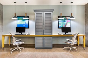 A modern office space with two desks, each with a computer monitor, keyboard, and phone. There are two office chairs, a large cabinet with louvered doors, and pendant lights above the desks. - Home2 Suites by Hilton Dallas Addison (Addison)