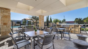 Un área de patio al aire libre con pilares de piedra, mesas y sillas de metal, y vistas a una piscina y edificios al fondo.