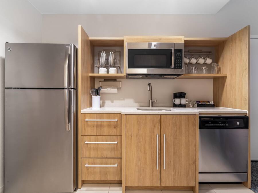 A compact kitchen with a stainless steel refrigerator, a microwave, a dishwasher, wooden cabinets, and a countertop with a sink.