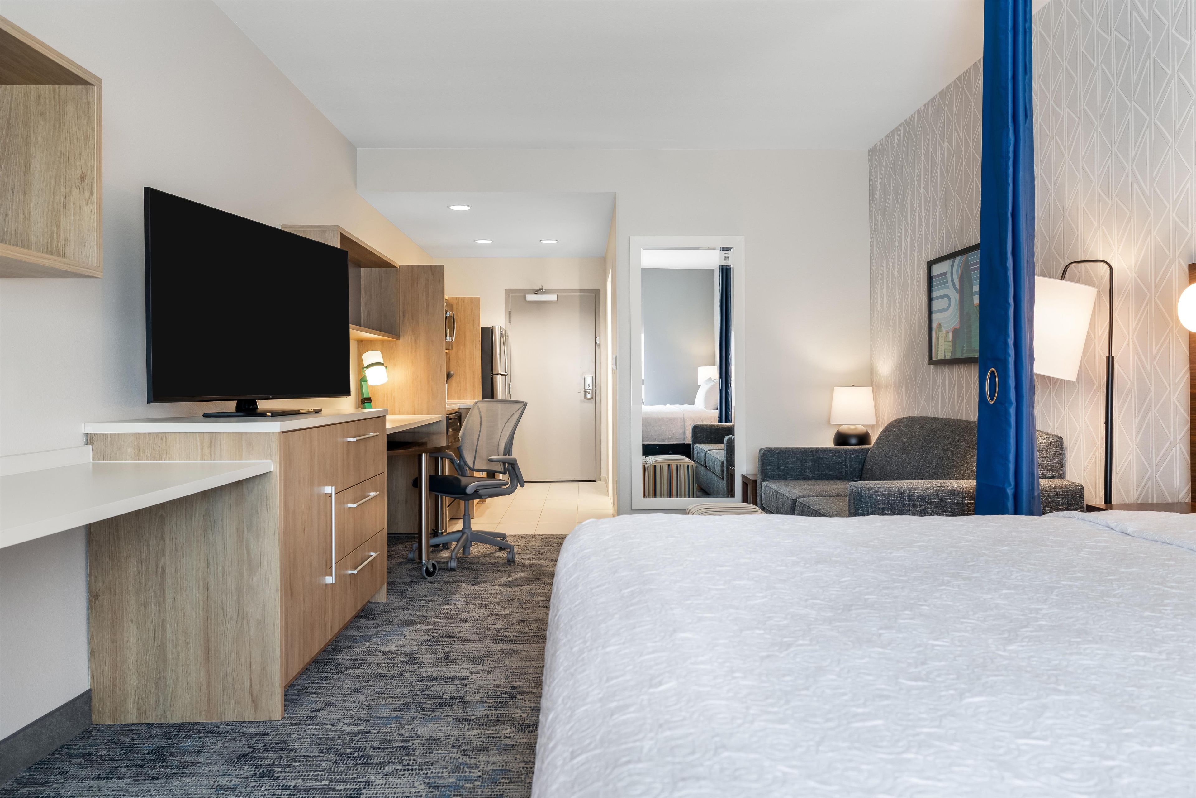 A hotel room with a bed, a desk with a flat-screen TV, a chair, a sofa, and a mirror.