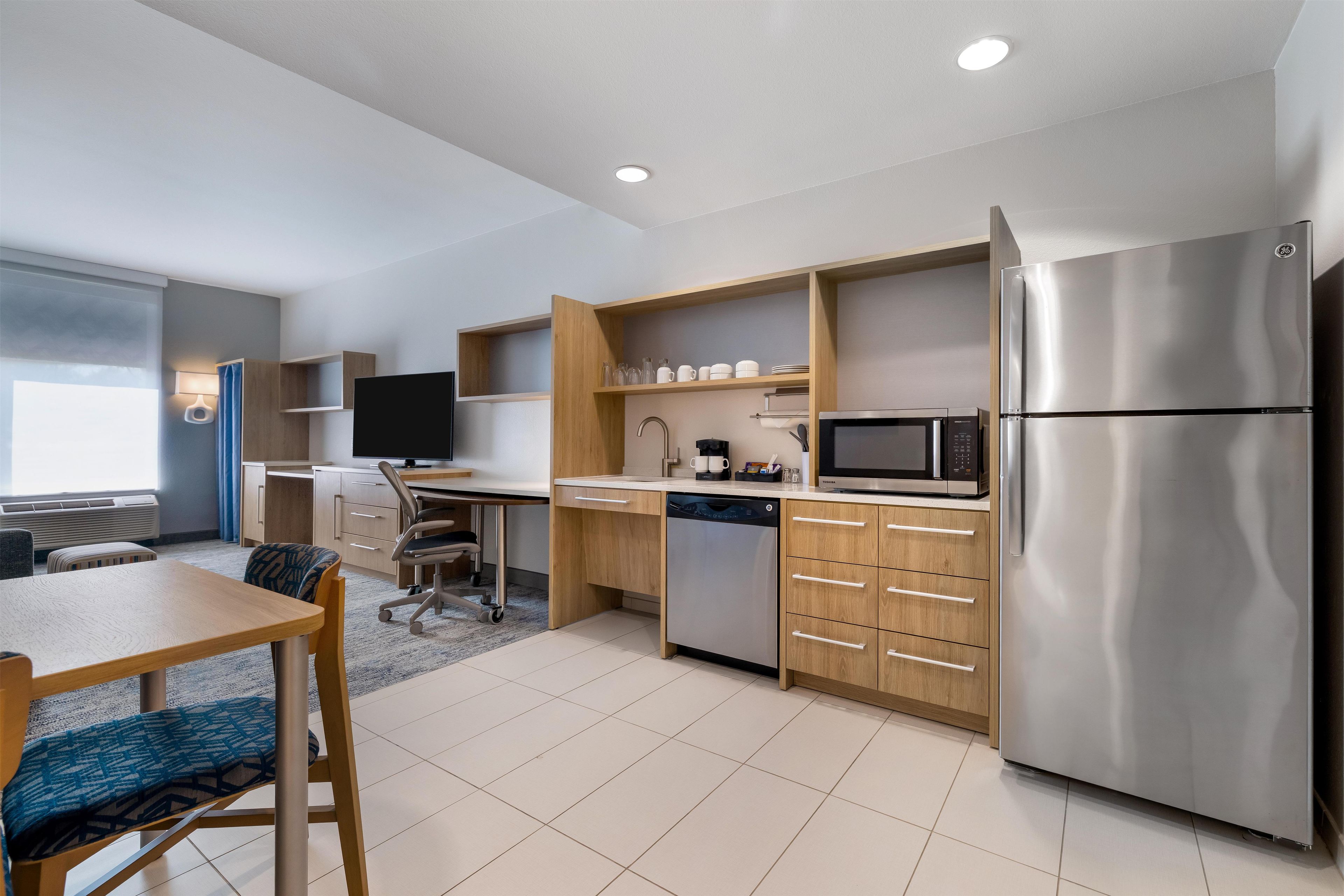 A modern kitchen with a stainless steel refrigerator, built-in microwave, and a dishwasher. There is a wooden dining table with chairs, and a television mounted on the wall.
