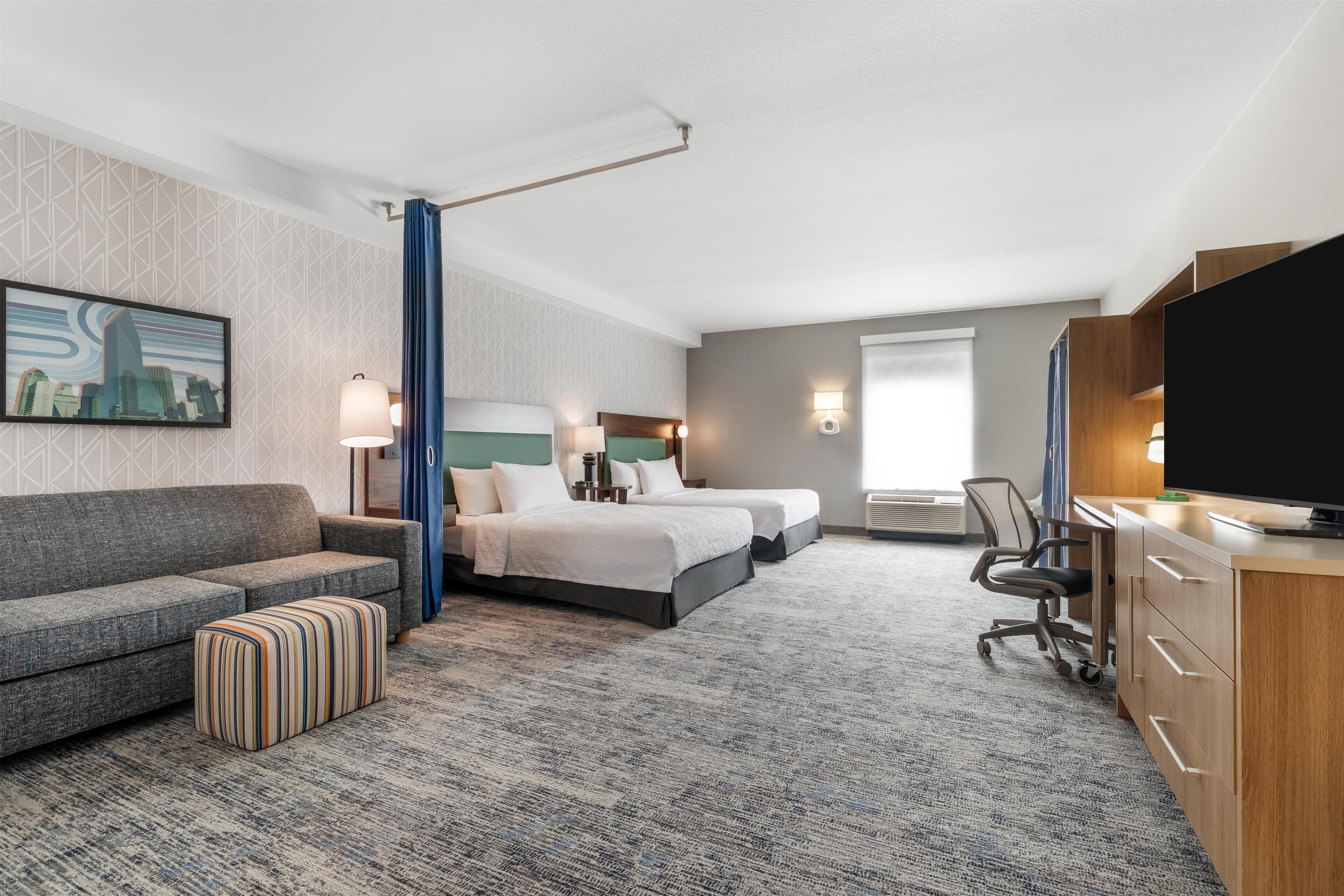 A hotel room with a gray sofa, two beds, a desk, a television, and a cityscape painting on the wall.
