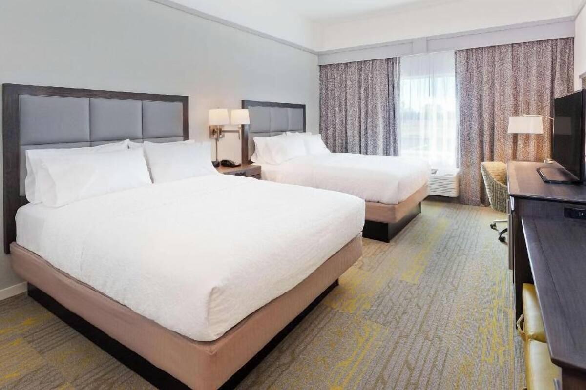 A hotel room with two beds, a desk, a TV, and a window with curtains.