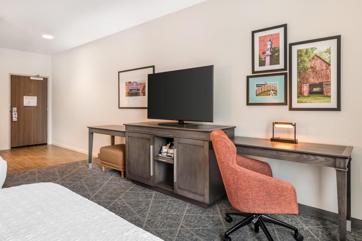 A hotel room with a bed, a desk with a TV, a chair, and framed pictures on the wall.