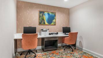 Una sala de conferencias con dos sillas de oficina, un escritorio con dos monitores, una impresora y un cuadro grande en la pared.