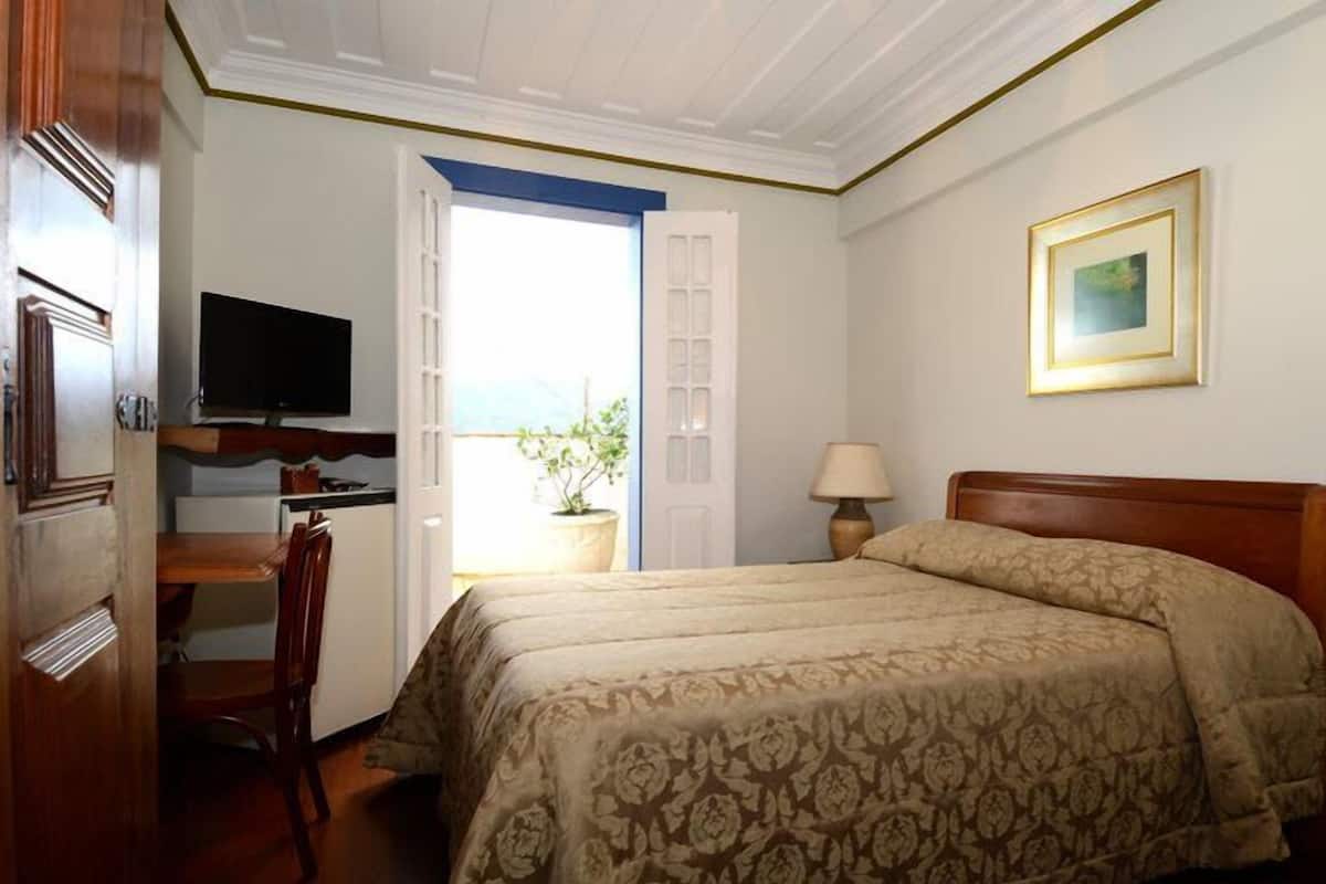Habitación de hotel con una cama, un escritorio con televisor, una silla, una lámpara y una puerta que da a un balcón.