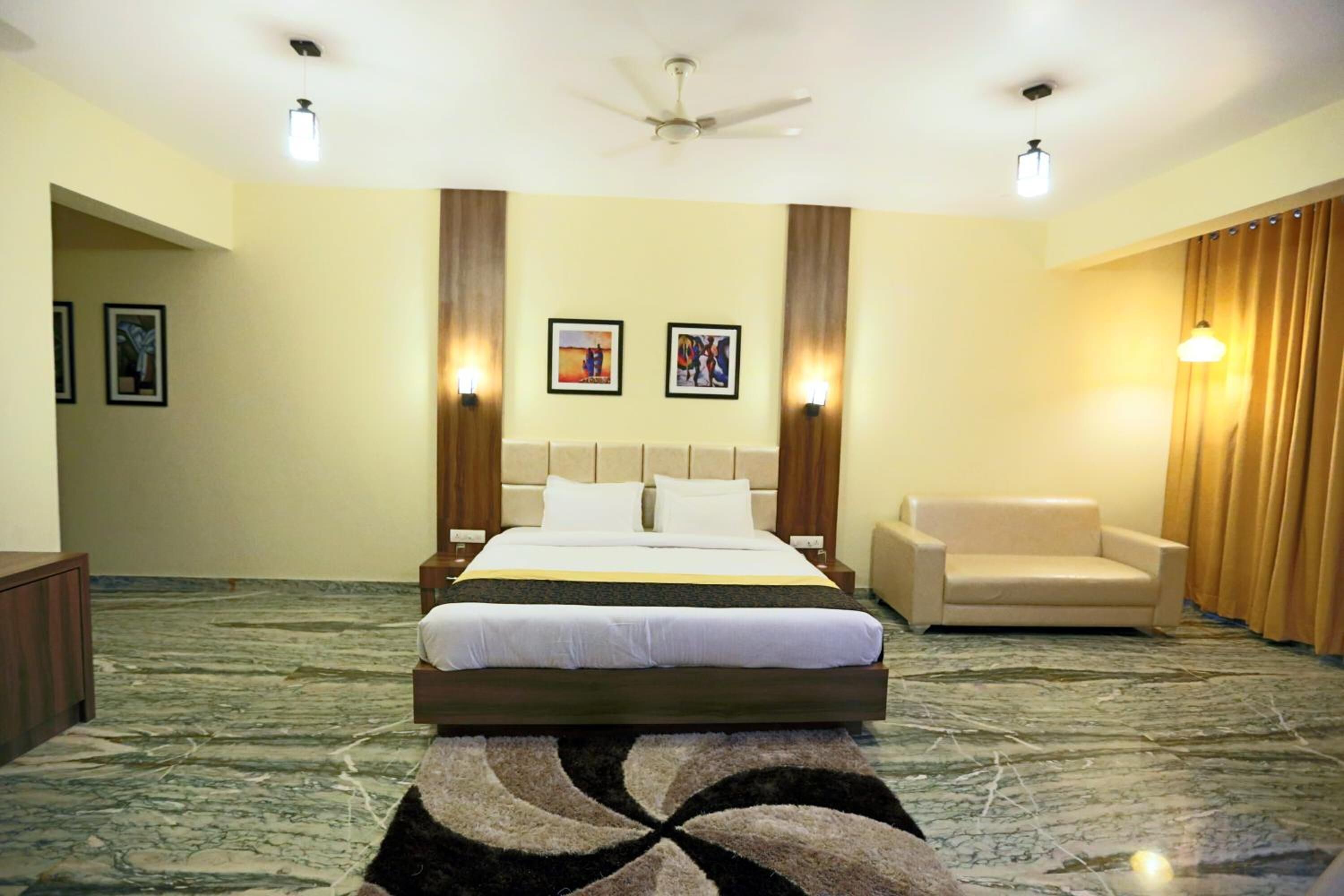 Une chambre d’hôtel avec un lit, un canapé, un ventilateur de plafond et des œuvres d’art encadrées accrochées aux murs.