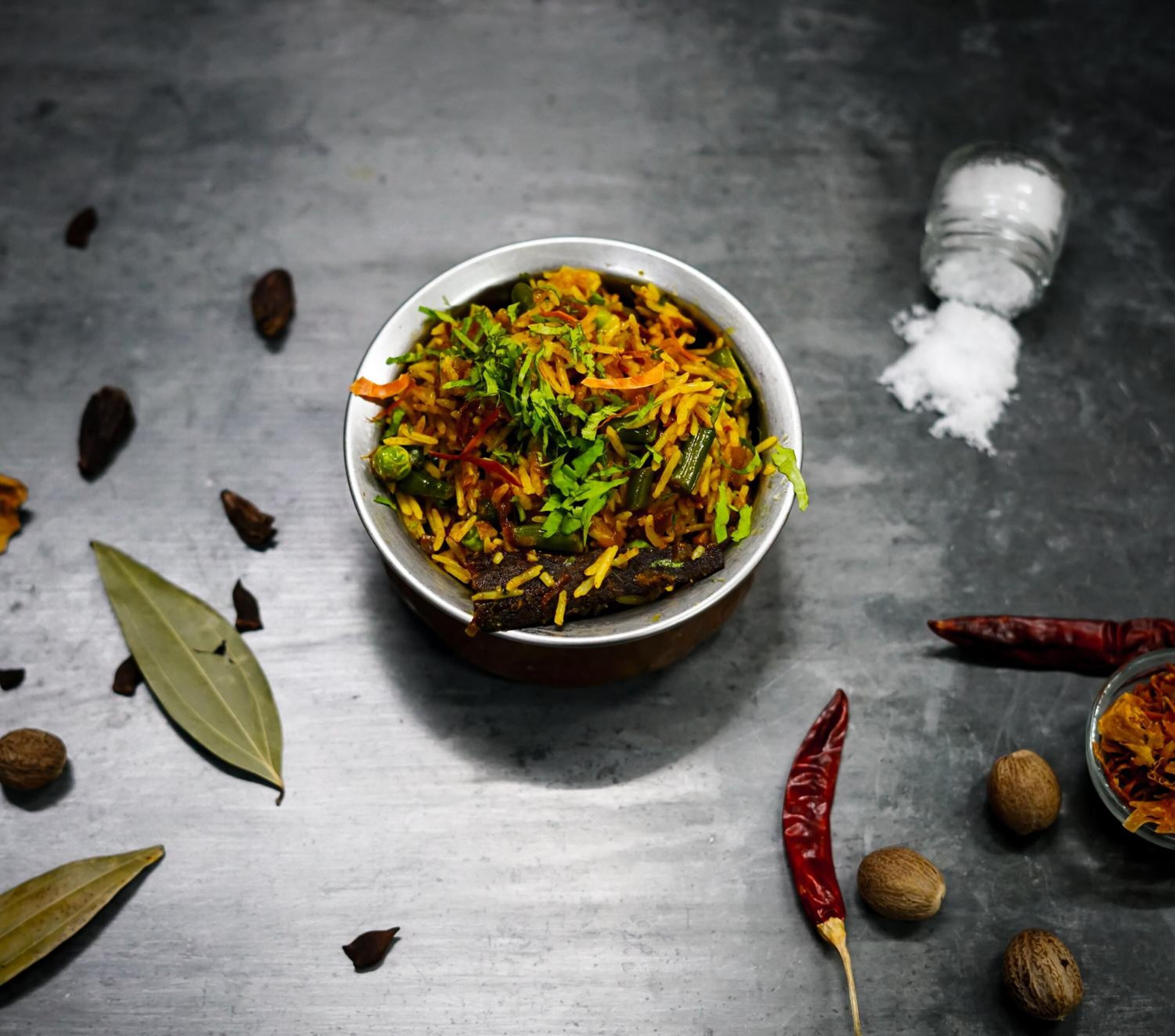A bowl of rice dish with vegetables and herbs, surrounded by spices and a salt shaker on a metallic surface.