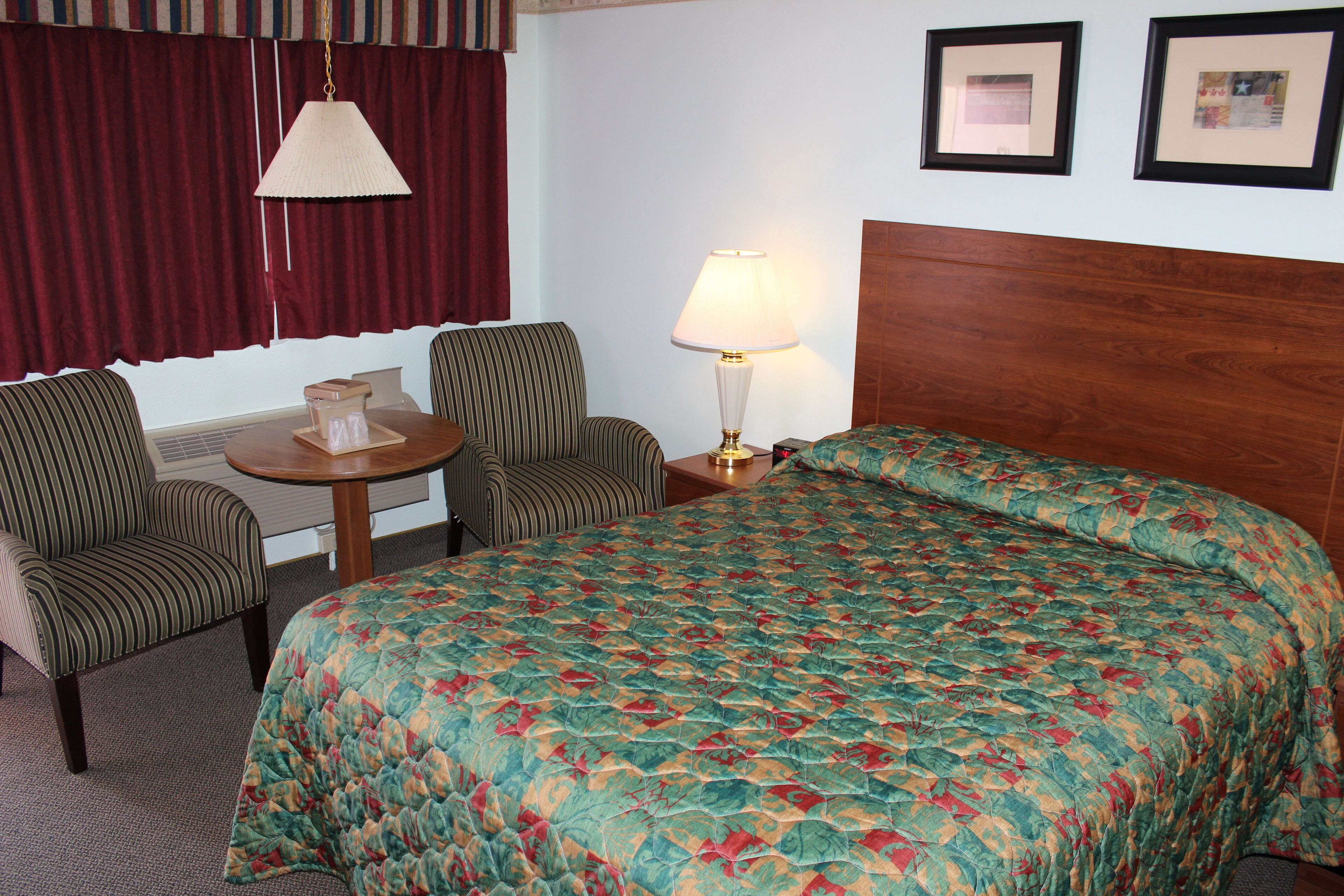 A hotel room with a bed, two chairs, a table, a lamp, and two framed pictures on the wall.