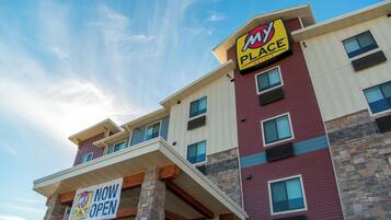 Un hotel de varios pisos con un cartel que dice 'NOW OPEN', con una fachada de piedra y madera, múltiples ventanas y un llamativo logo con la letra 'M'.
