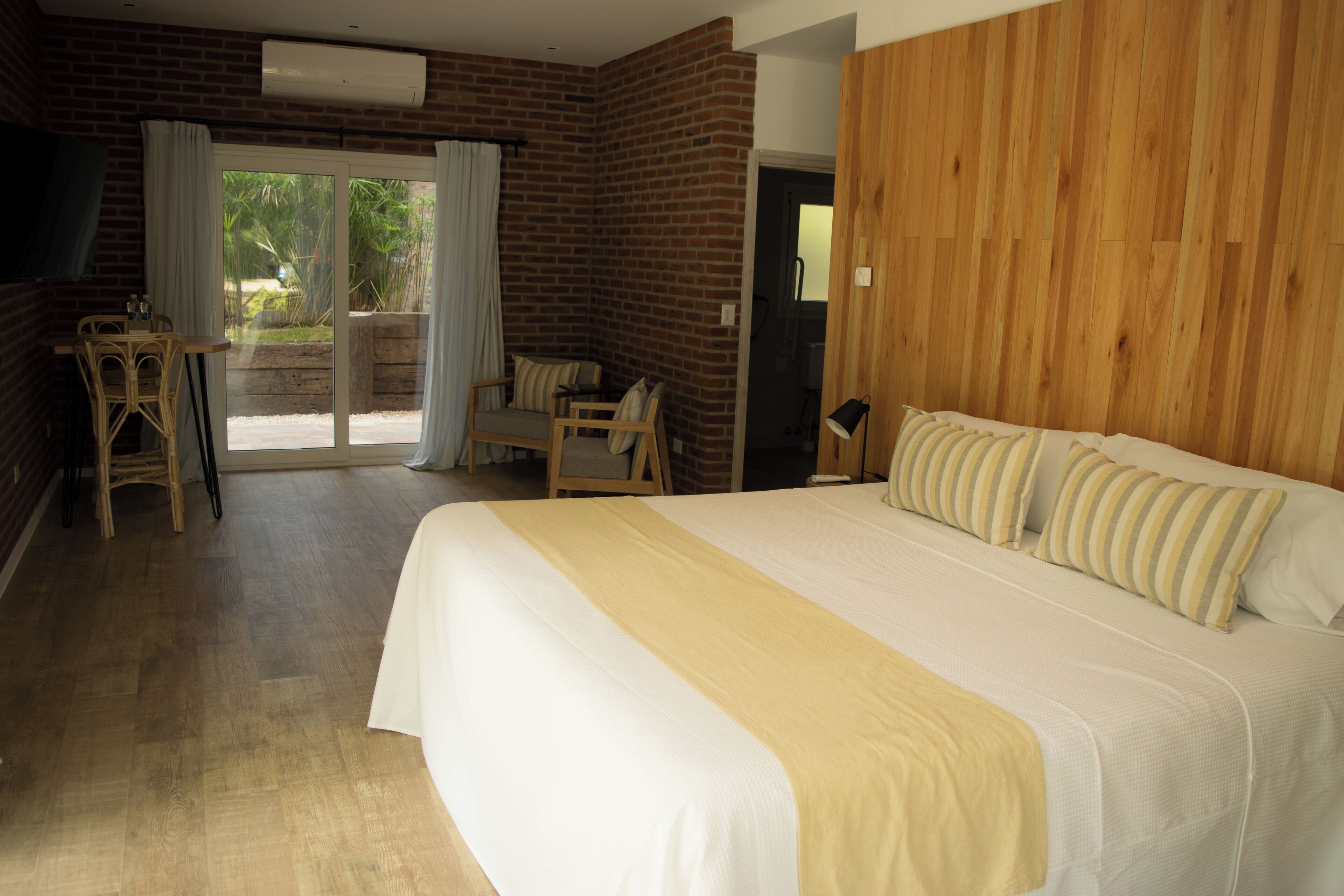 A hotel room with a large bed, a wooden wall, a balcony with chairs, and a TV.