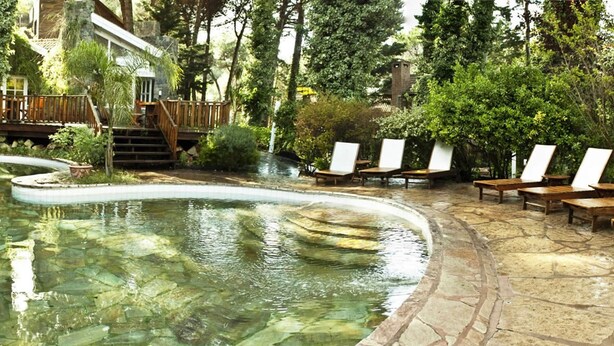 A pool area with a stone deck, wooden lounge chairs, and a wooden deck with stairs leading to a building. - Carilo Village Apart Hotel & Spa (Carilo)