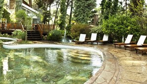 A pool area with a stone deck, wooden lounge chairs, and a wooden deck with stairs leading to a building.