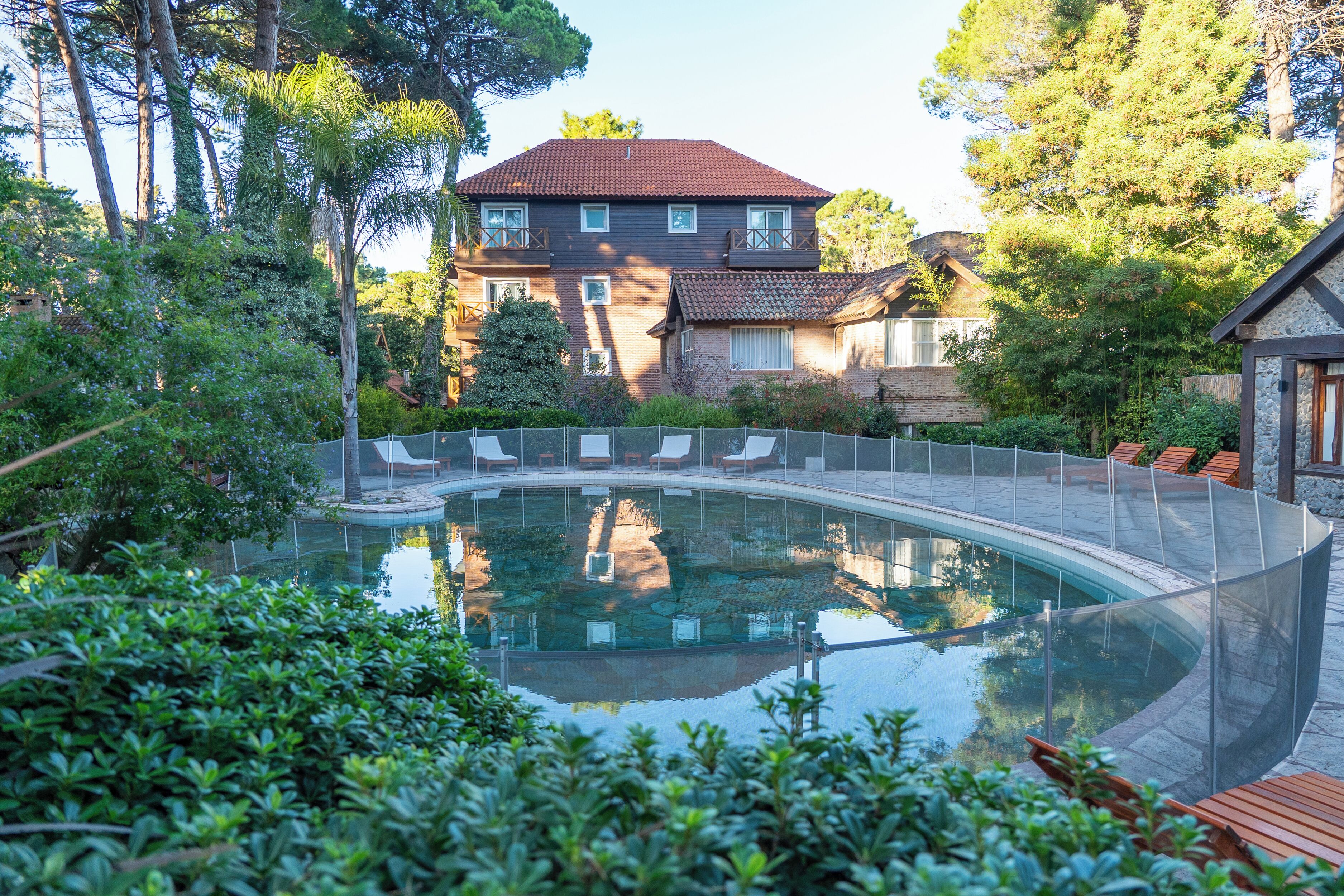 Una piscina con un cerramiento de vidrio curvo, rodeada de abundante vegetación y una casa de dos plantas al fondo.