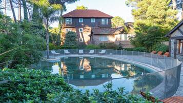 Una piscina con un cerramiento de vidrio curvo, rodeada de abundante vegetación y una casa de dos plantas al fondo.