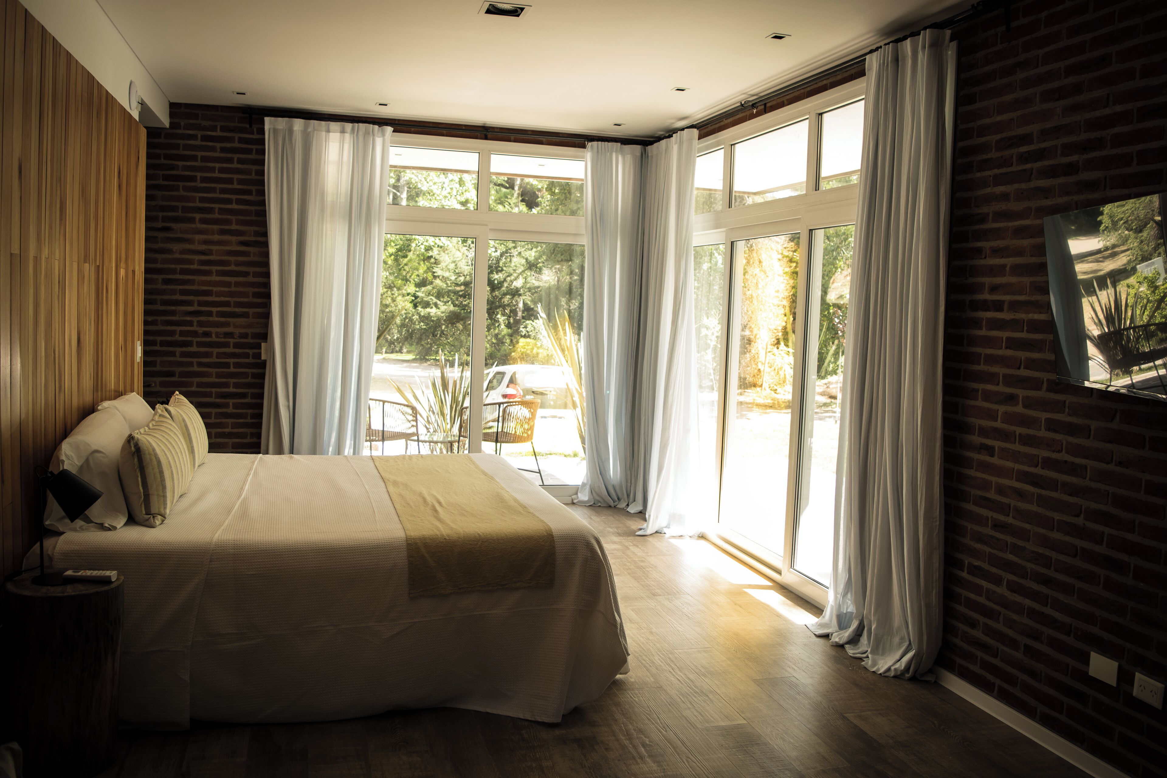 A modern bedroom with a large bed, a wooden headboard, and a view of the outdoors through large windows.