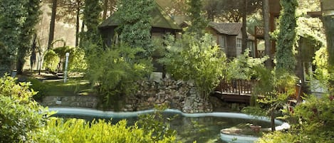 A serene garden with a pond, stone wall, and wooden structures surrounded by lush greenery.
